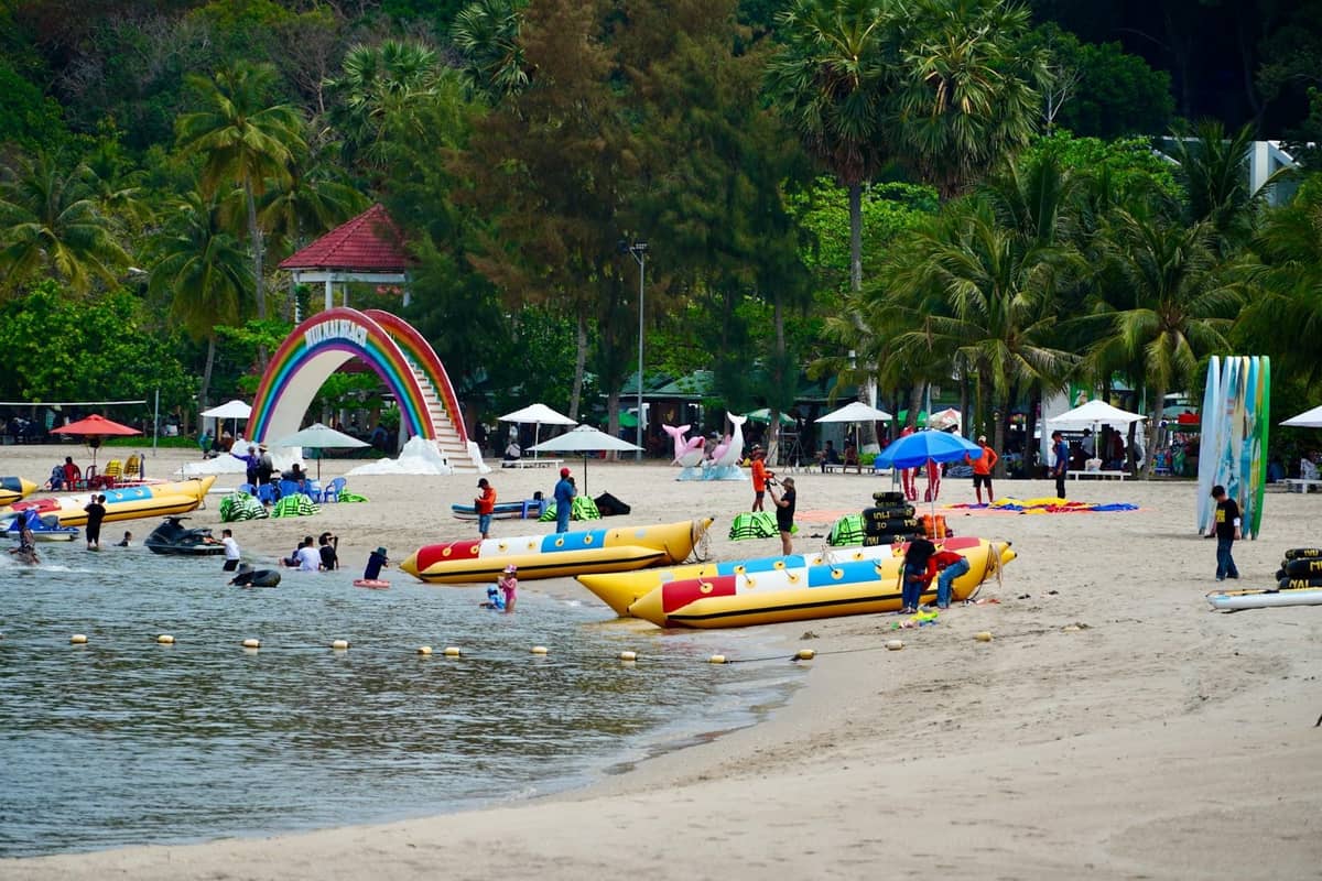 Families and children enjoy water activities and games on a sunny Pattaya beach with colorful banana boats and a large rainbow arch.