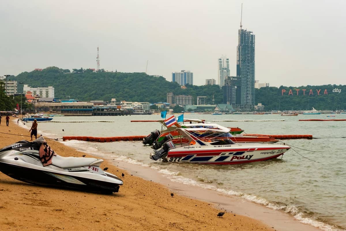 Jet skis and speedboats on a sandy Pattaya beach with city skyline and Pattaya City sign in the background.