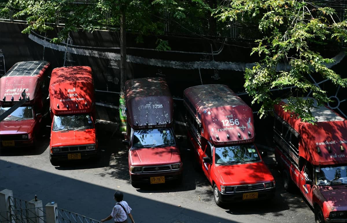 Several red songthaew taxis parked in Pattaya, shaded by trees, with a pedestrian walking nearby.