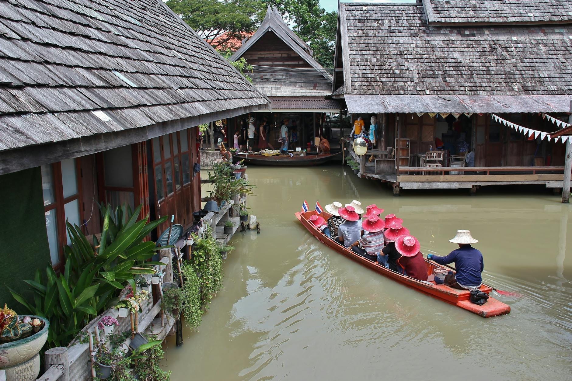 Barche di legno condotte da guide con turisti in cappelli colorati, che scivolano lungo canali stretti circondati da rustiche case su palafitte in stile thai al Mercato Galleggiante di Pattaya.