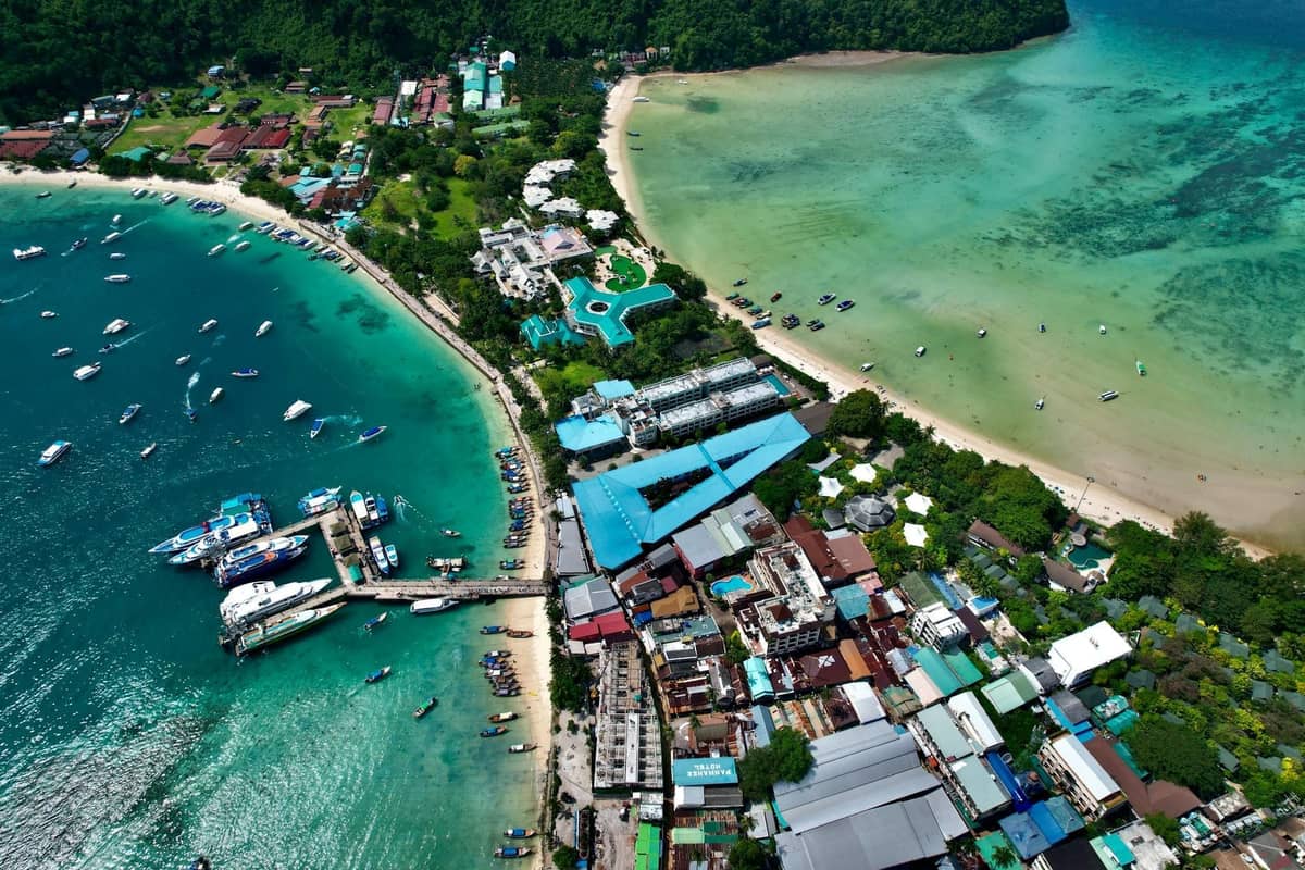 Aerial view of a busy pier, boats, and beach on Koh Larn Island with clear green water and nearby buildings, vibrant and full of activity.