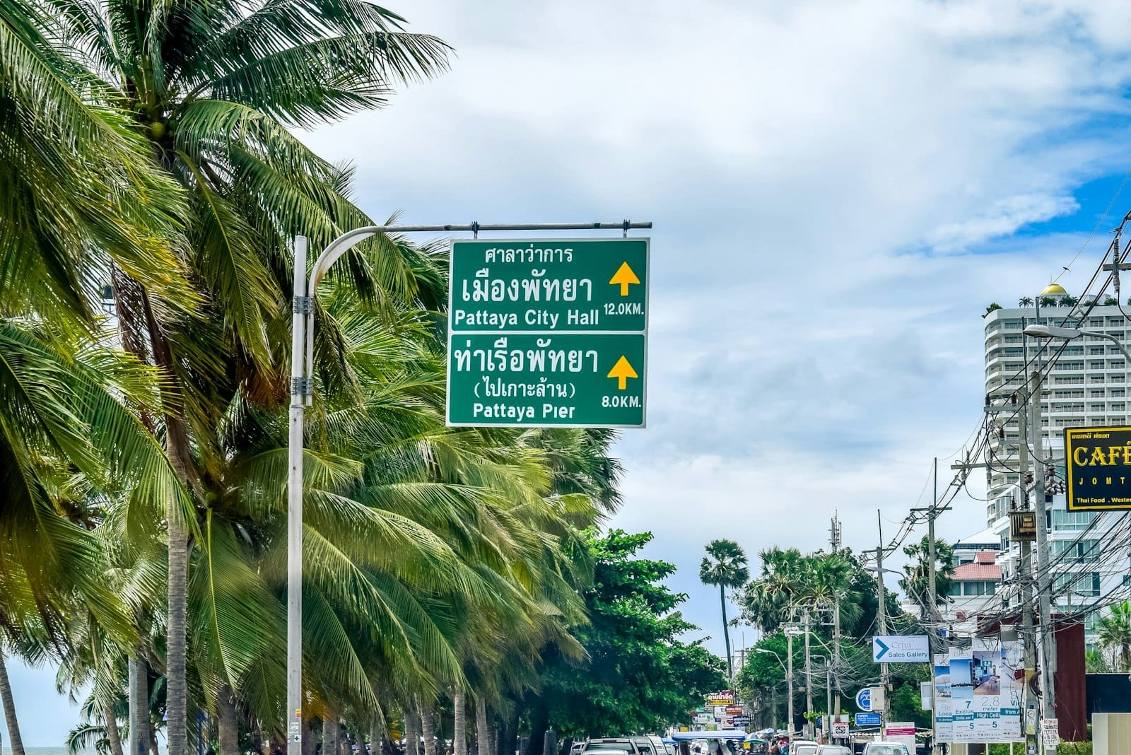 Placas de rua verdes para o Pattaya City Hall e o Pattaya Pier com palmeiras, edifícios e tráfego ao longo de uma ensolarada estrada costeira.