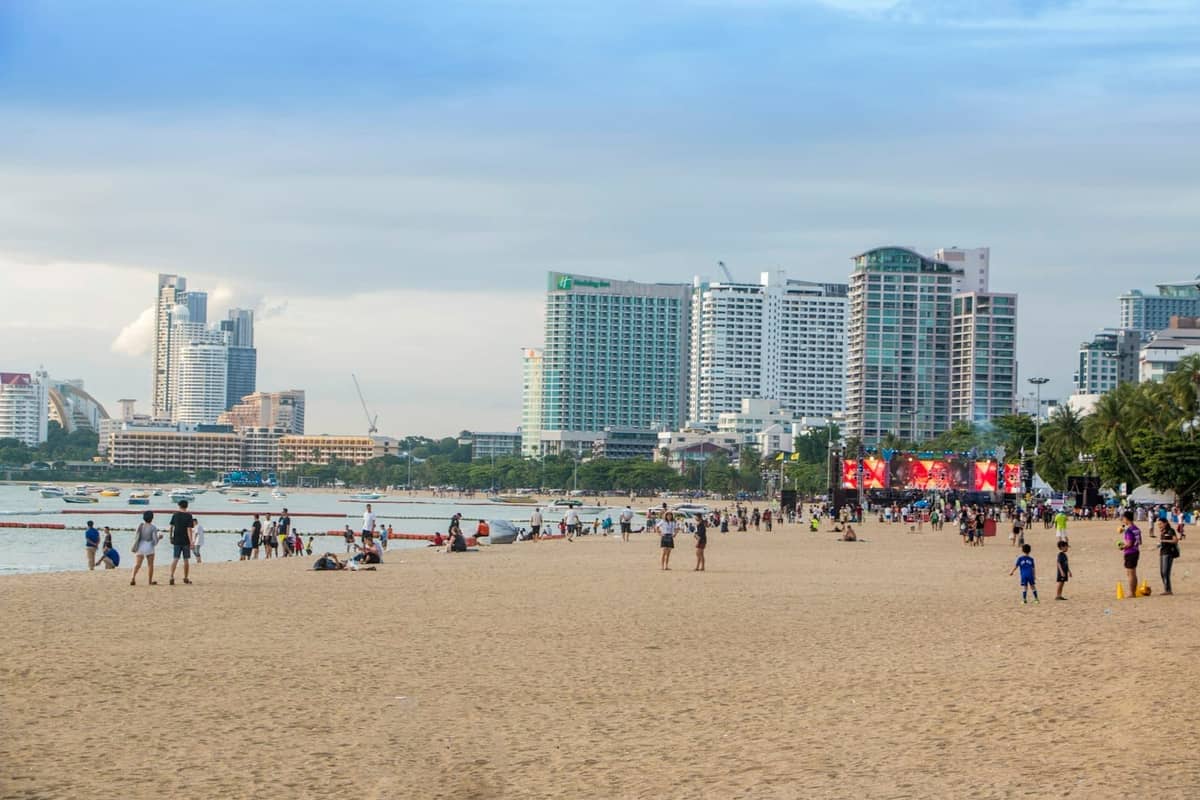 People relaxing and walking along Pattaya Beach with city buildings and the sea in the background on a bright, clear day.