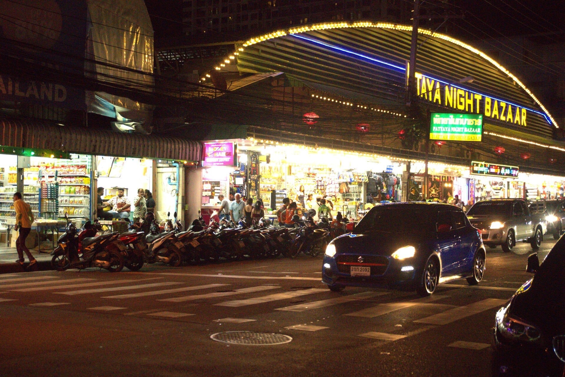 Luci brillanti illuminano l'ingresso del Pattaya Night Bazaar di notte, con motociclette e auto parcheggiate all'esterno e acquirenti visibili all'interno.