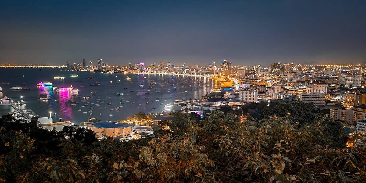 Nighttime cityscape of Pattaya with illuminated buildings, neon lights, and boats in the bay, showing the city's vibrant nightlife from an elevated viewpoint.