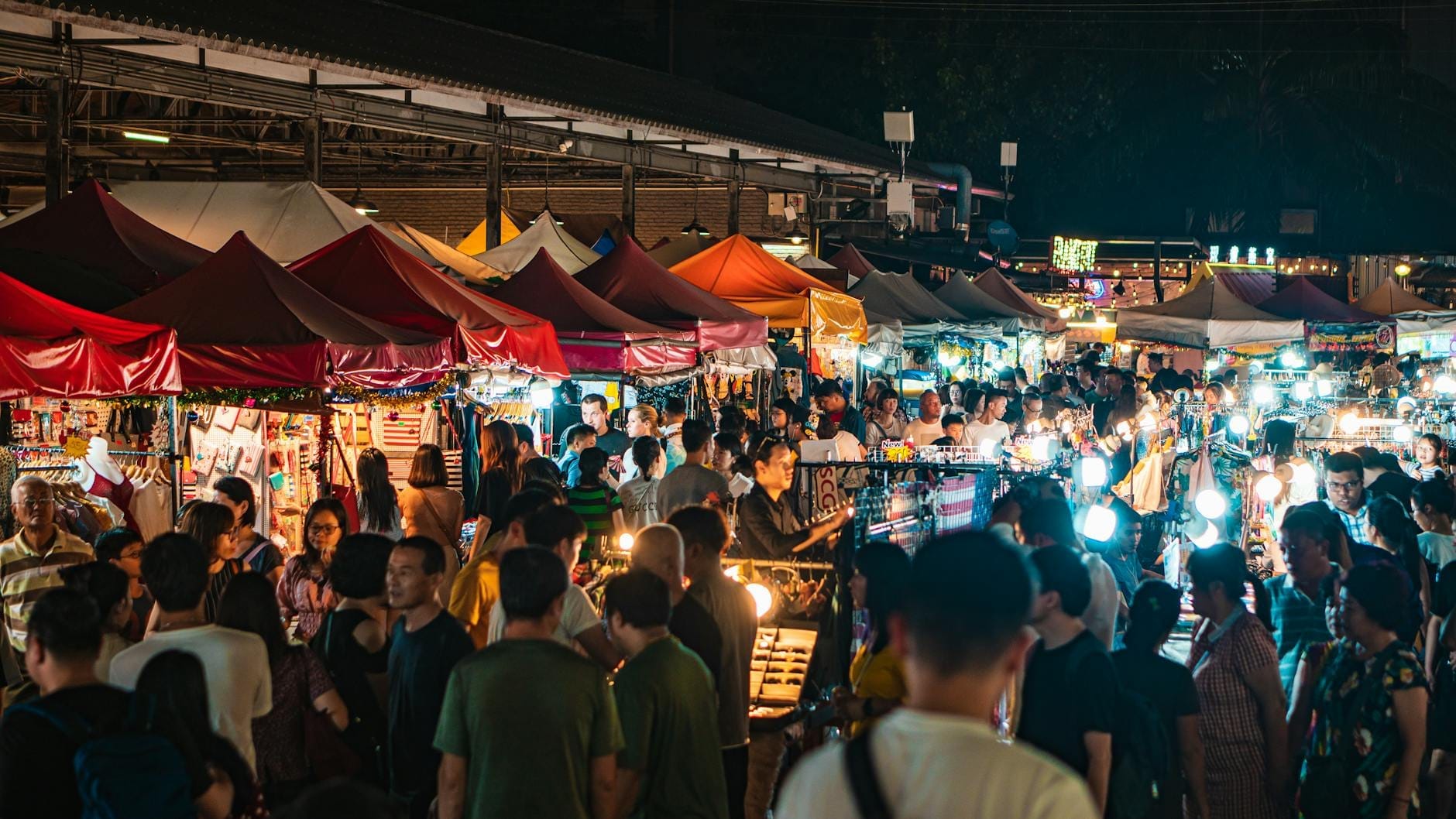 Le folle esplorano il Pattaya Park Night Market di notte, curiosando tra le bancarelle sotto tende colorate con luci vivaci e venditori locali visibili.