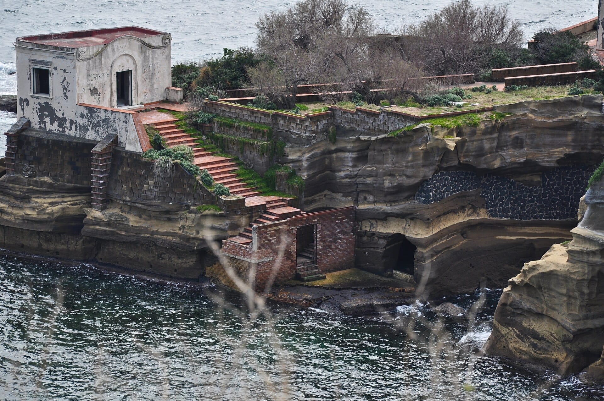 Gradini in pietra consumati e un'antica villa al Parco Archeologico del Pausilypon si ergono sopra la costa rocciosa e il mare blu.