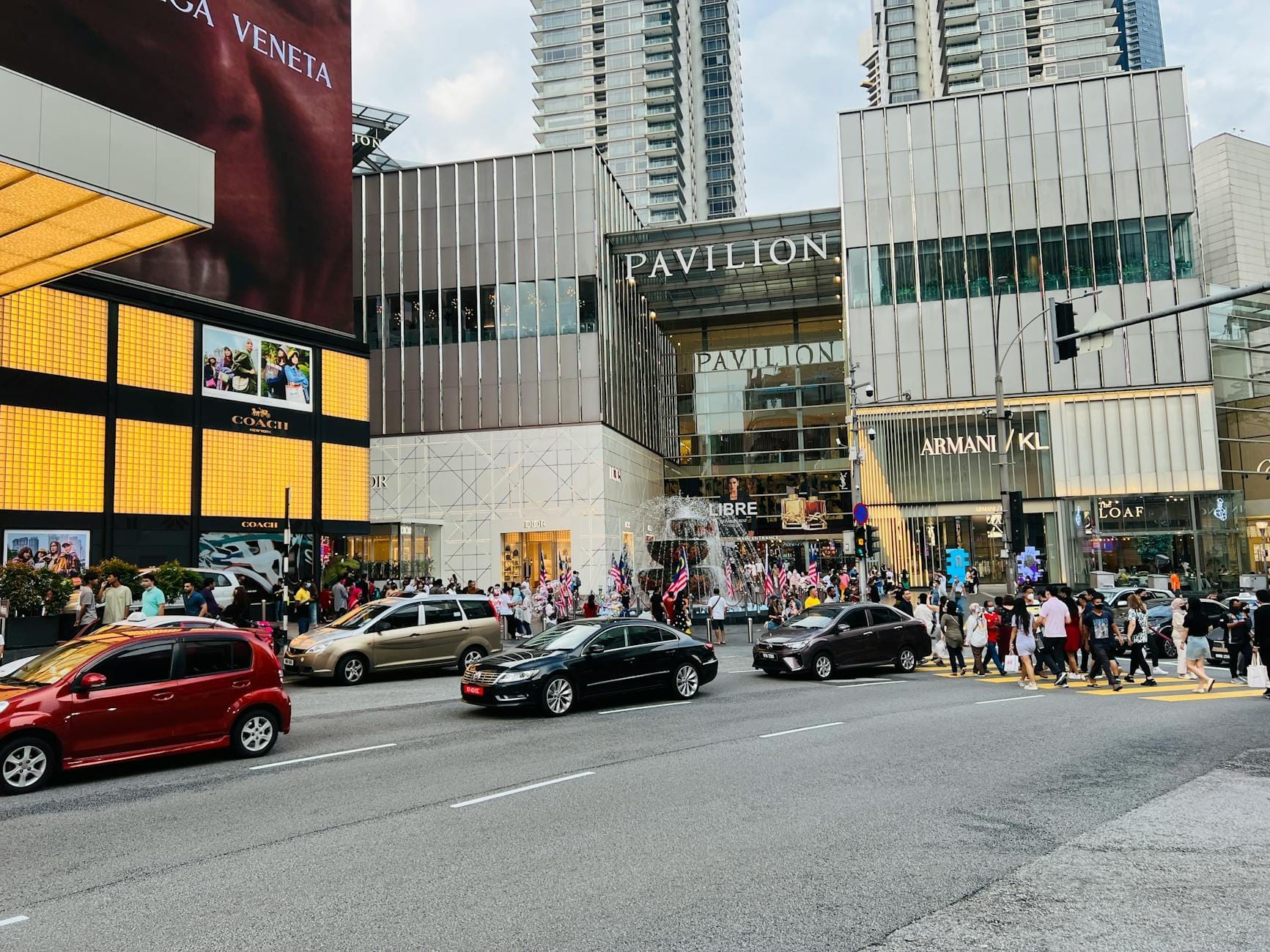 Street view of Pavilion Kuala Lumpur’s main entrance with luxury brand stores, busy crosswalk, modern buildings, and a crowd of shoppers.