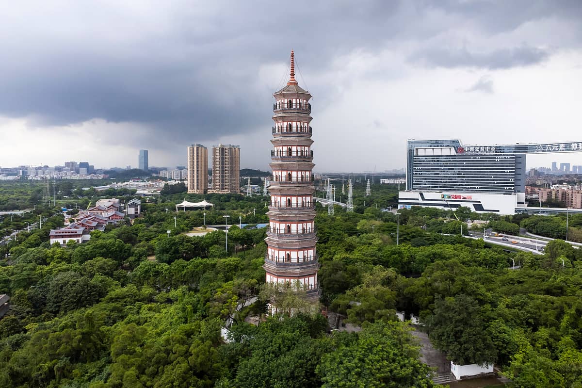 Aerial view of Pazhou Pagoda, a tall octagonal brick tower surrounded by lush greenery, with the Guangzhou city skyline and modern buildings in the background.