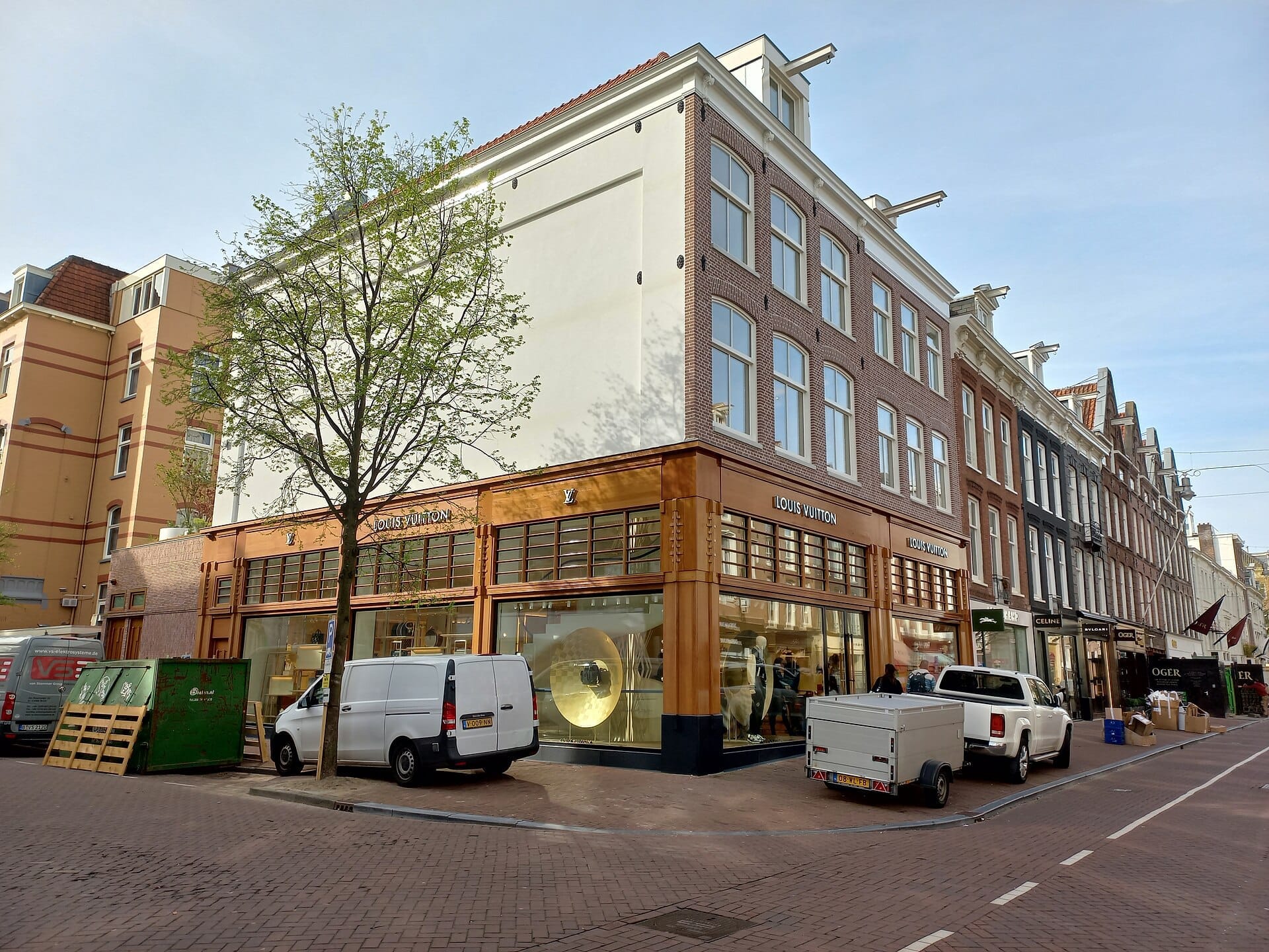 Fachadas modernas de marcas de lujo como Louis Vuitton bordean la PC Hooftstraat en Ámsterdam, con grandes escaparates, furgonetas aparcadas y un cielo azul despejado.