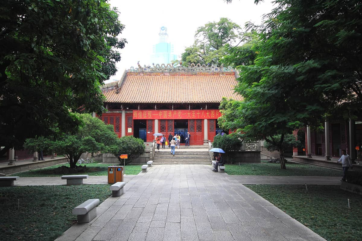 Front view of the historic Peasant Movement Training Institute in Guangzhou, showing red doors, decorative roof, and visitors walking toward the entrance.