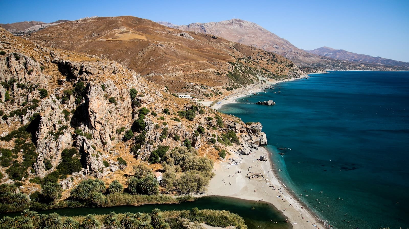 มุมมองทางอากาศของ Pefkos Beach แสดงให้เห็นทรายสีทอง ทะเลสีฟ้าเทอร์ควอยซ์ หน้าผาหิน และนักอาบแดดที่กระจายอยู่ใต้ท้องฟ้าสีฟ้าใส