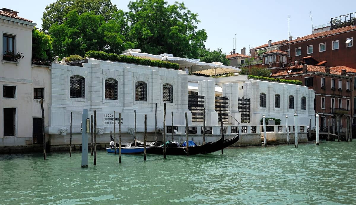 Die weiße Palazzo-Fassade der Peggy Guggenheim Collection am Canal Grande in Venedig, mit schwarzen Gondeln im Wasser und Grünpflanzen über dem Gebäude.