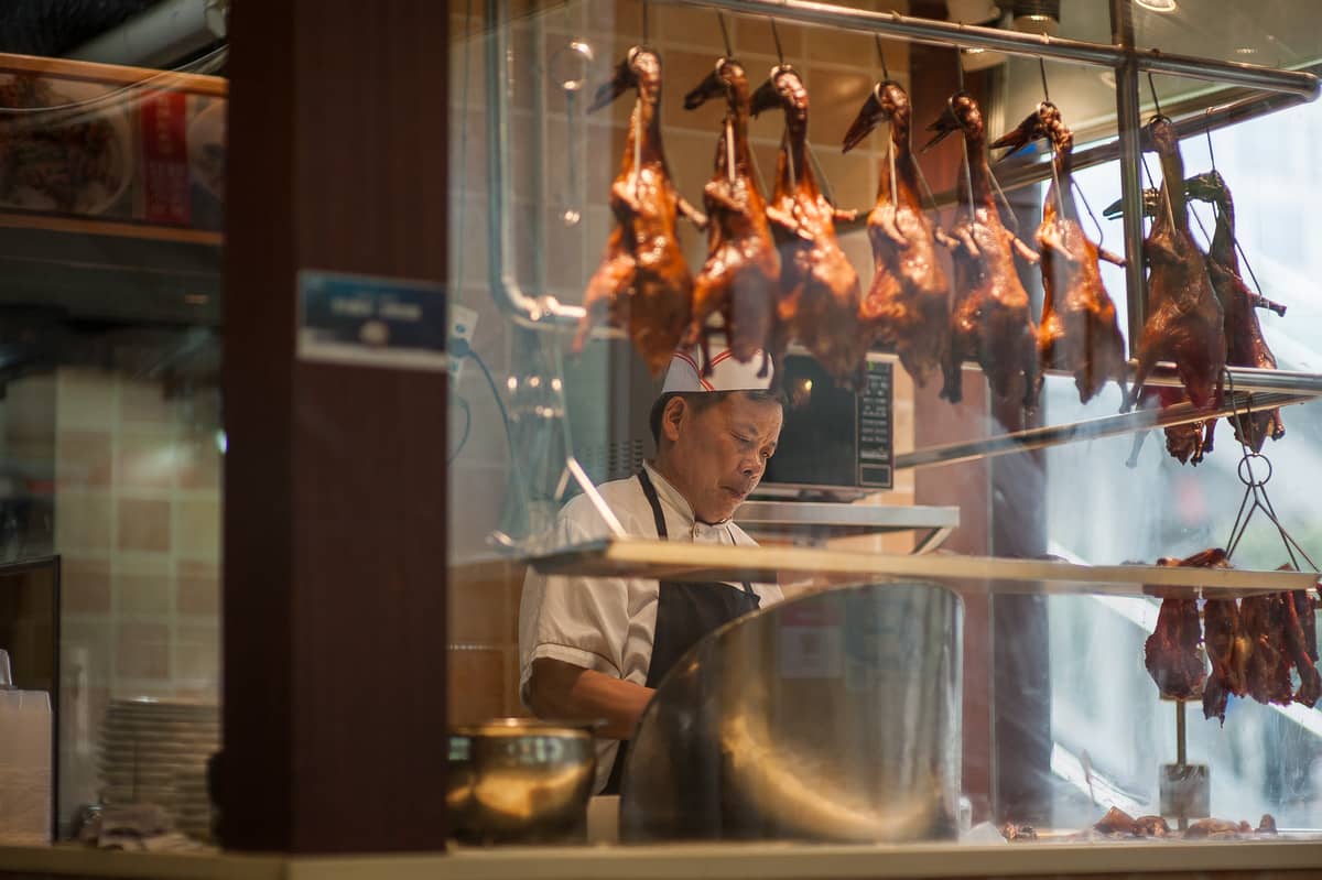 Chef behind glass counter slicing roasted ducks hanging above in a traditional Chinese kitchen, with shiny lacquered poultry visible.