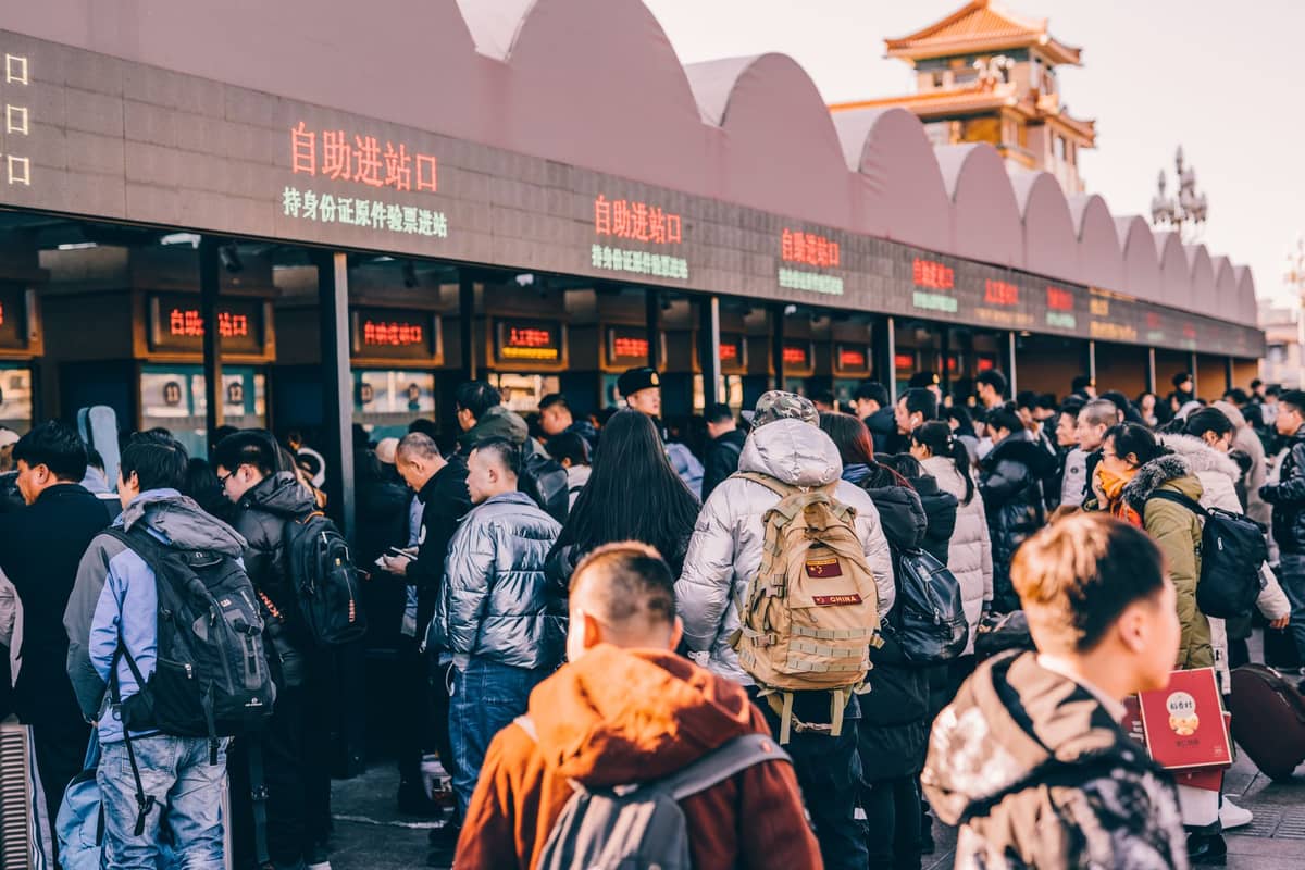Large crowd of people queueing outdoors at ticket counters with Chinese signage near a traditional style Beijing building.