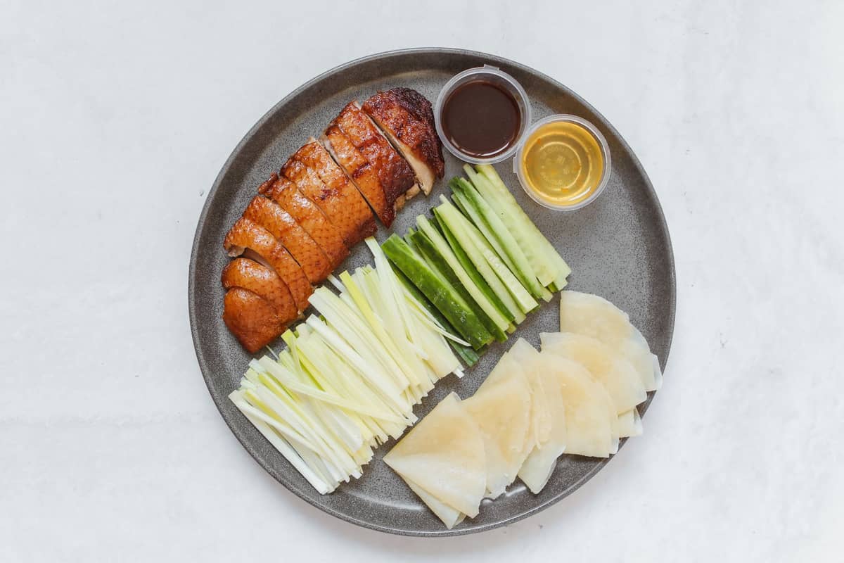 A plate with sliced Peking duck, cucumber, spring onion, wheat pancakes, and sauces arranged neatly on a gray plate.
