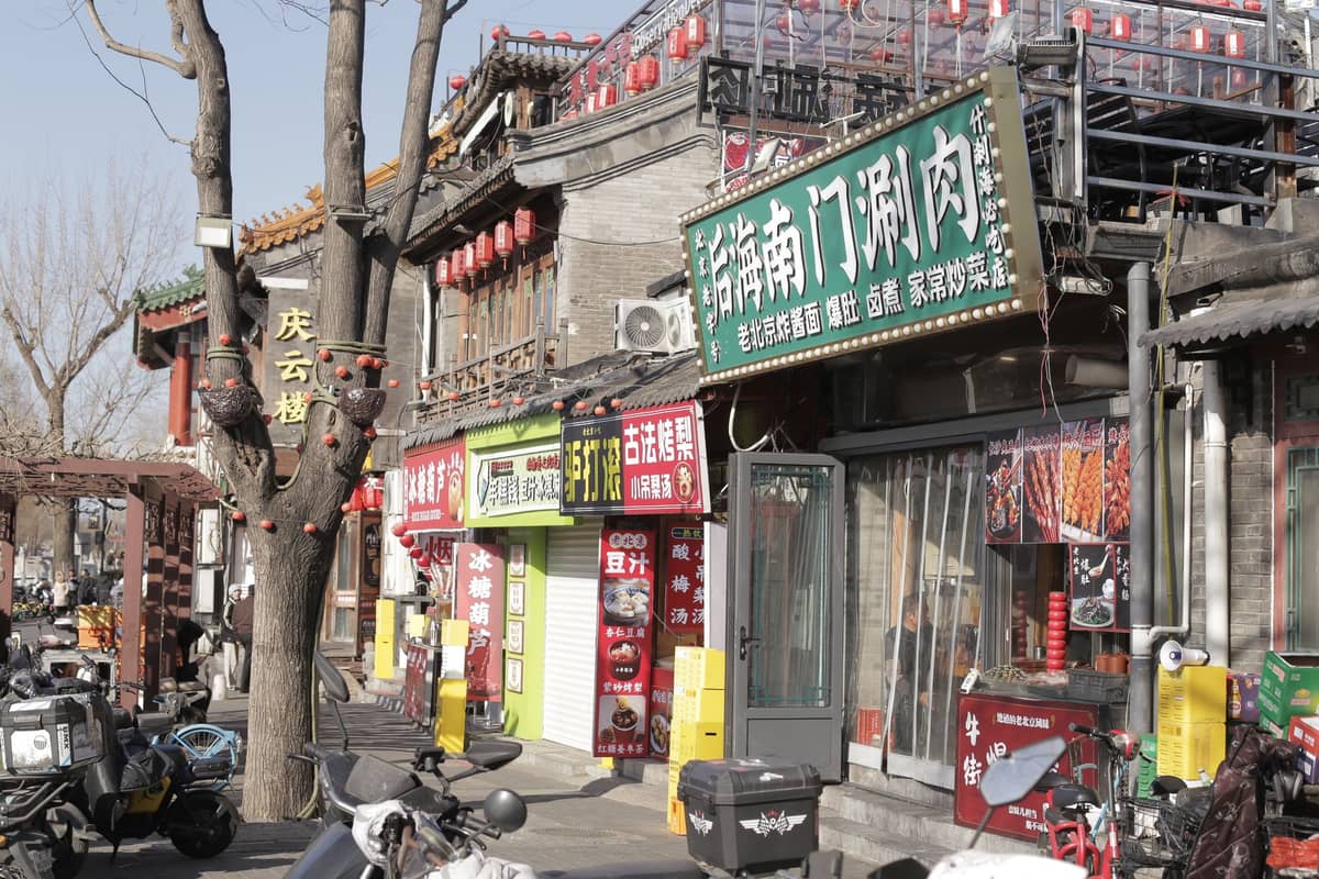 A row of bustling restaurants with colorful signs, scooters parked outside, and hints of traditional Beijing hutong architecture.