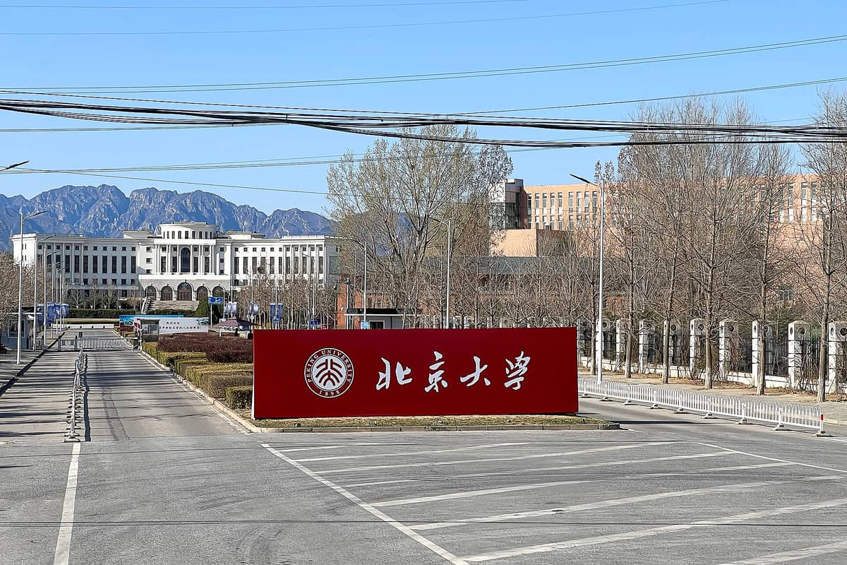 Weitwinkelaufnahme des Haupteingangs der Peking-Universität mit einem großen roten Schild mit dem Universitätsnamen, Campusgebäuden und Bergen im Hintergrund.
