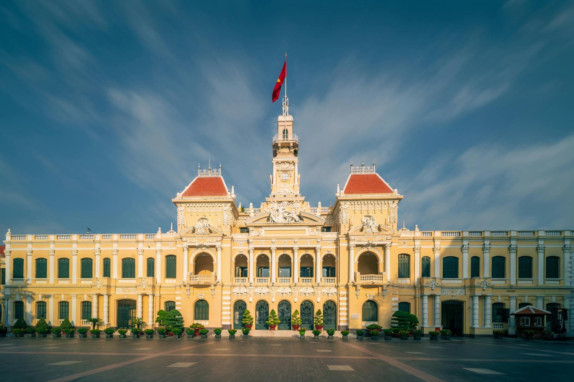 Vista frontal y amplia del Edificio del Comité Popular en Ho Chi Minh con su fachada colonial amarillo pálido, techo rojo y bandera vietnamita bajo un cielo azul intenso.