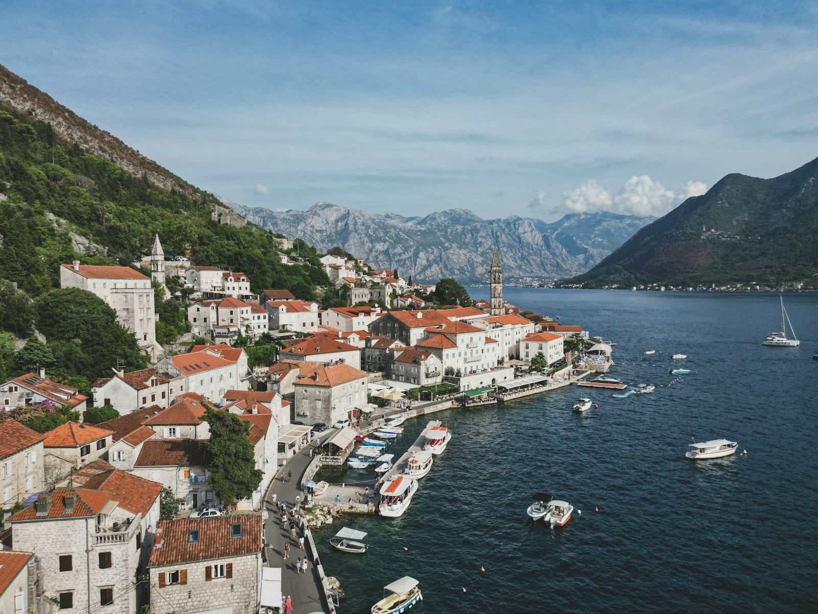 วิว Perast ริมน้ำแบบพาโนรามา ตึกบาโรกหลังคาแดงเรื่อยตามชายฝั่ง มีเรือจอดตลอดแนว ด้านหลังเป็นภูเขาสูงที่มองลงมายังอ่าวโคเตอร์