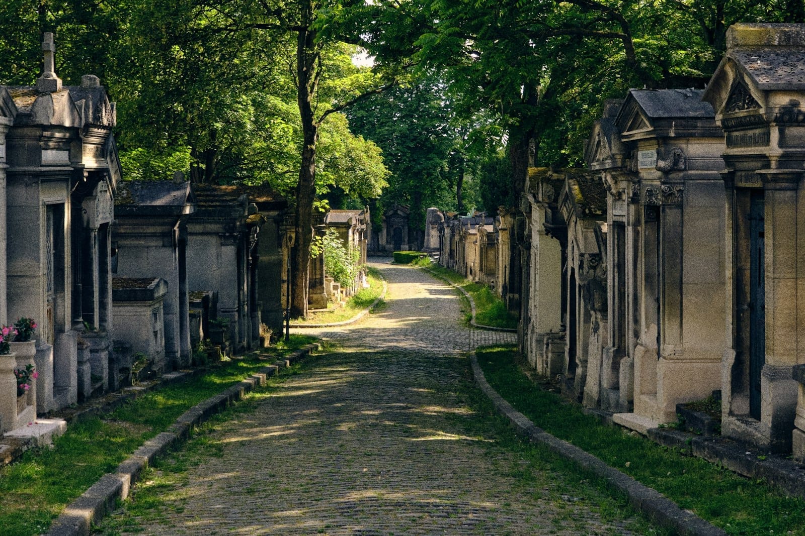 Sentiero in sampietrini illuminato dal sole che si snoda tra tombe elaborate e mausolei sotto alberi frondosi al cimitero del Père Lachaise a Parigi, evocando pace e storia.