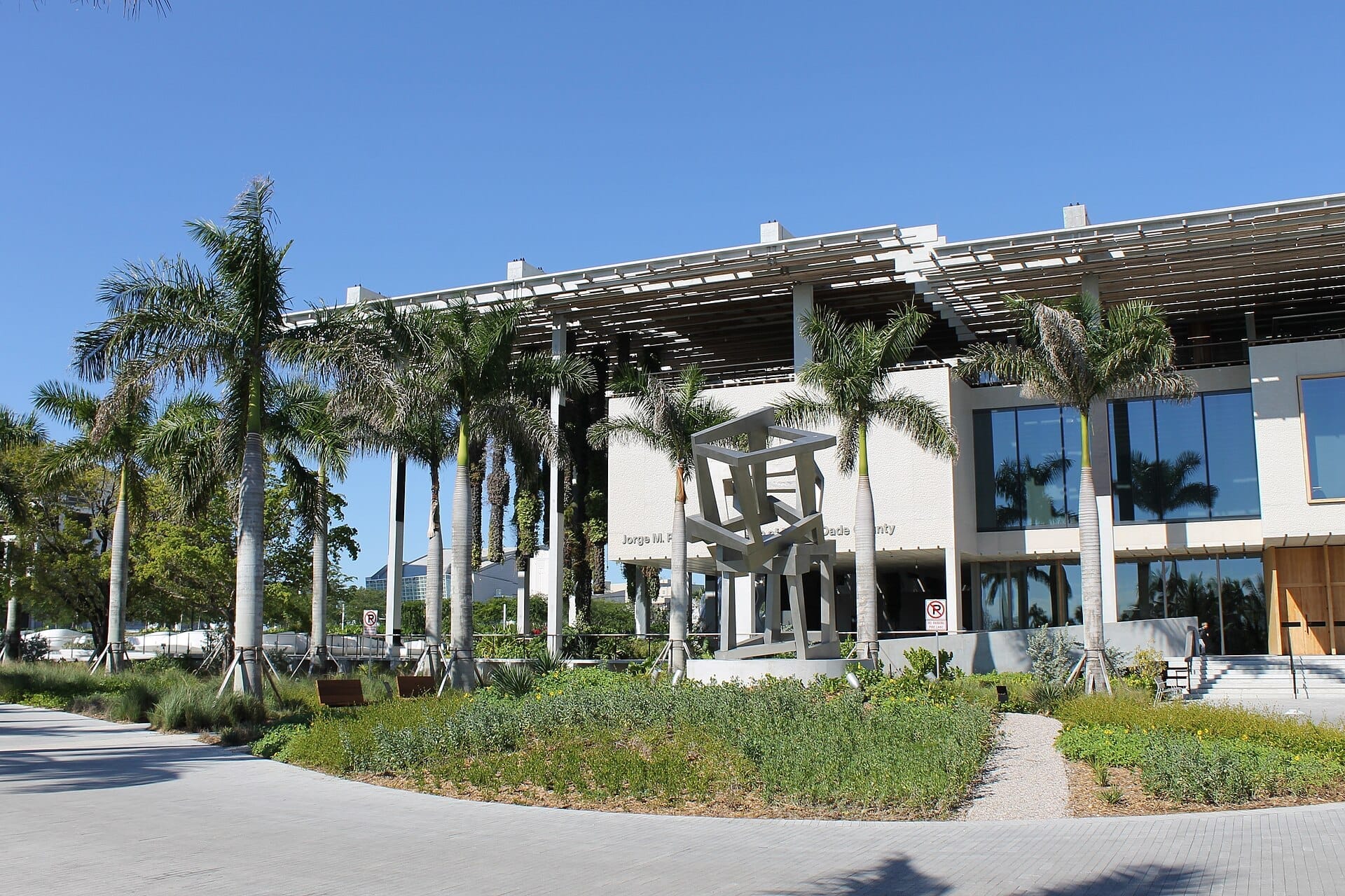 Vista frontale del Pérez Art Museum Miami con palme, architettura moderna, finestre in vetro e una scultura metallica geometrica sotto un cielo azzurro limpido.