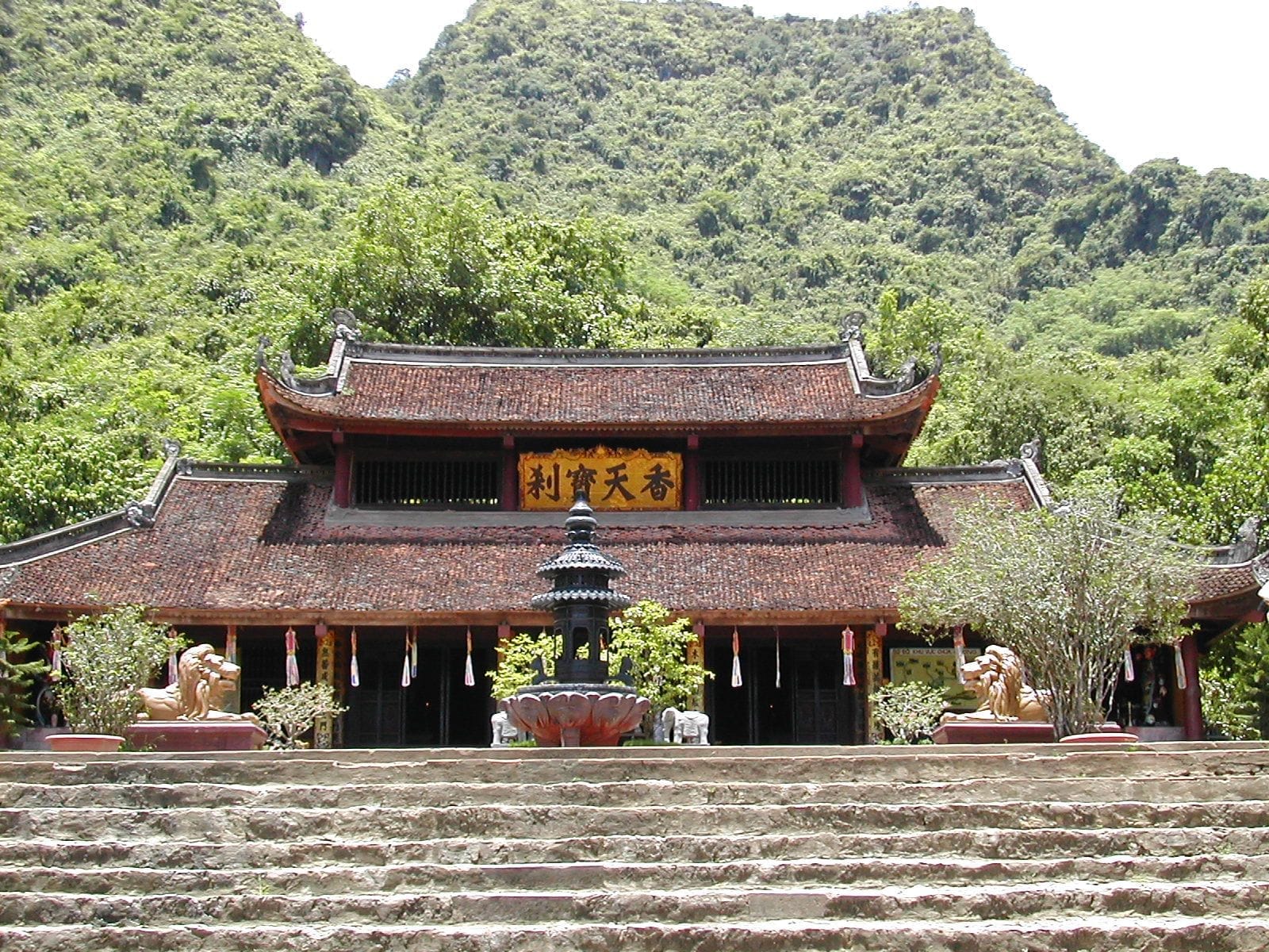 Main entrance of Perfume Pagoda with traditional red-tiled roof, stone steps, lion statues, and lush green mountains in the background.