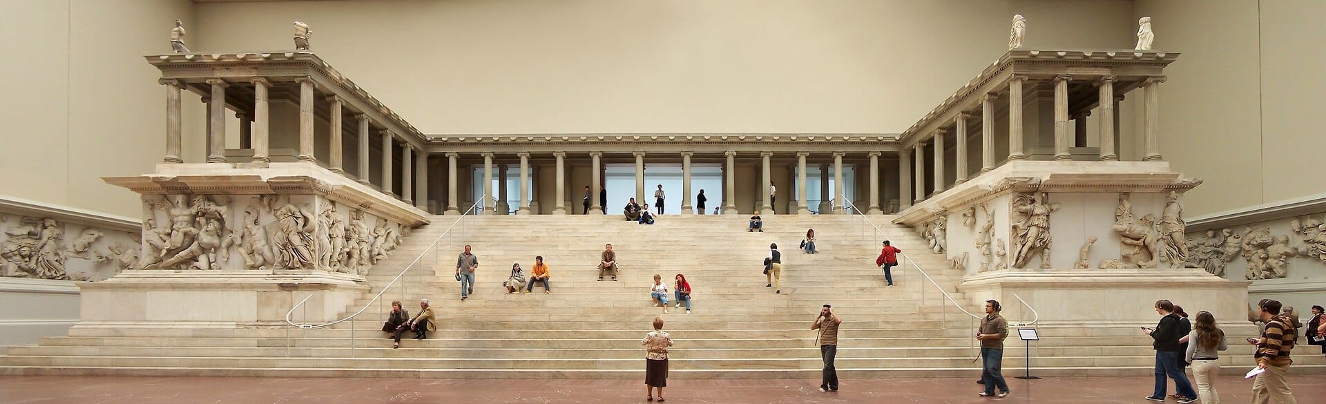 Il monumentale Altare di Pergamo all'interno del Pergamon Museum di Berlino, con visitatori che esplorano i grandi gradini di marmo e gli intricati fregi antichi sotto una soffusa illuminazione interna.