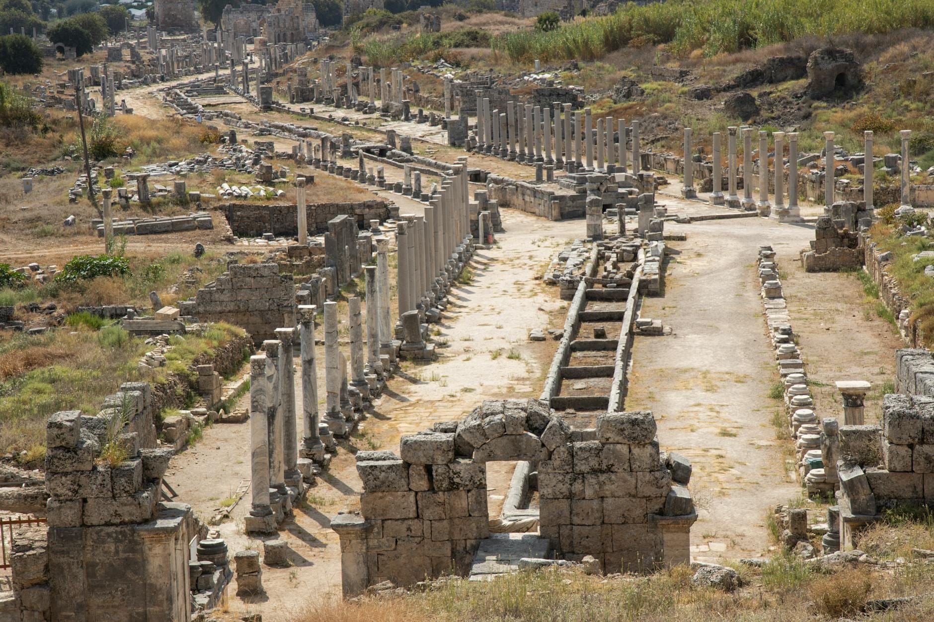 Panoramica della città antica di Perge con la lunga strada romana colonnata, le rovine in pietra e il suggestivo impianto archeologico sotto un cielo luminoso.