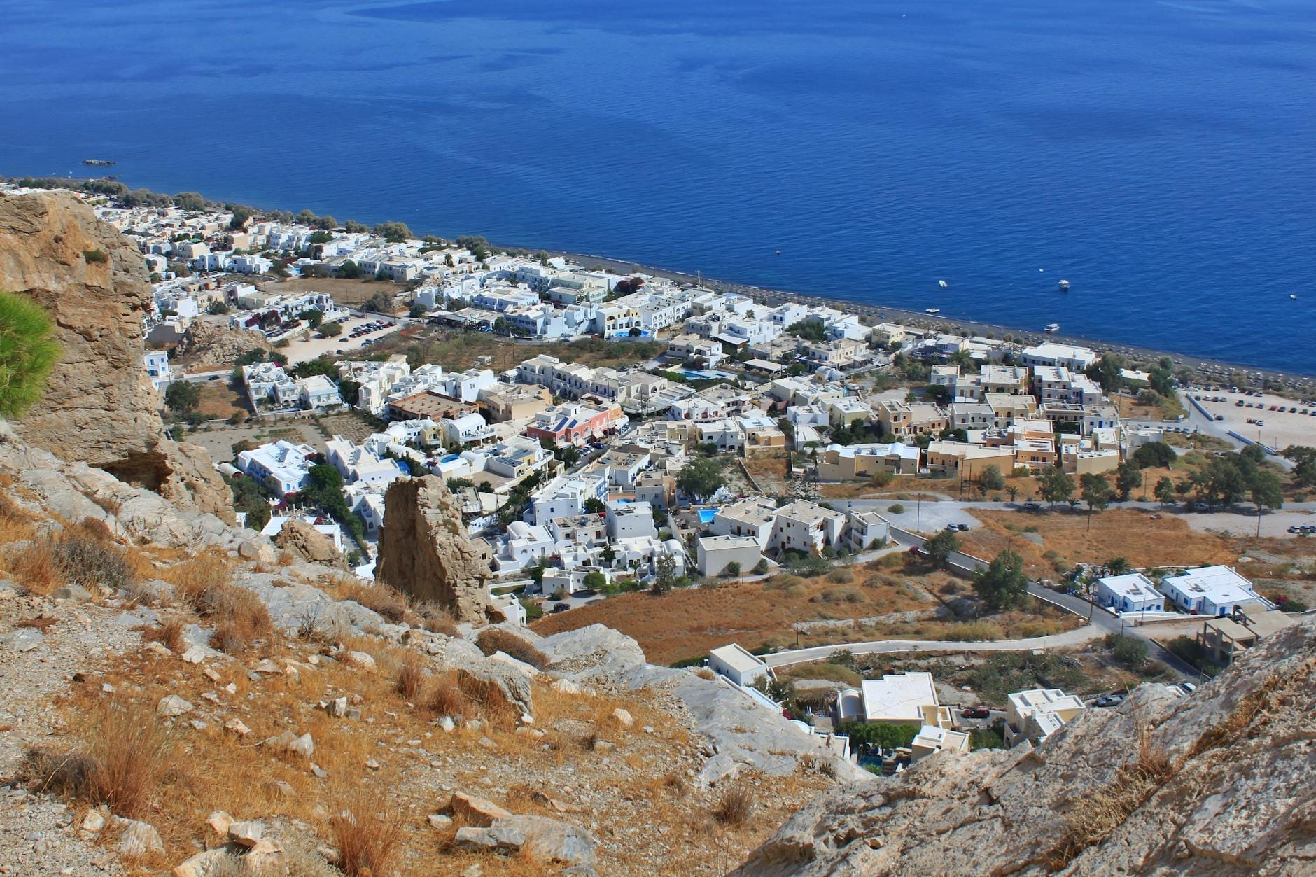 มุมมองทางอากาศของหมู่บ้าน Perissa บนเกาะ Santorini พร้อมอาคารทาสีขาว ลาดเขาหินแห้งแล้ง และทะเล Aegean สีน้ำเงินเข้มเบื้องหลังหาดทรายดำ