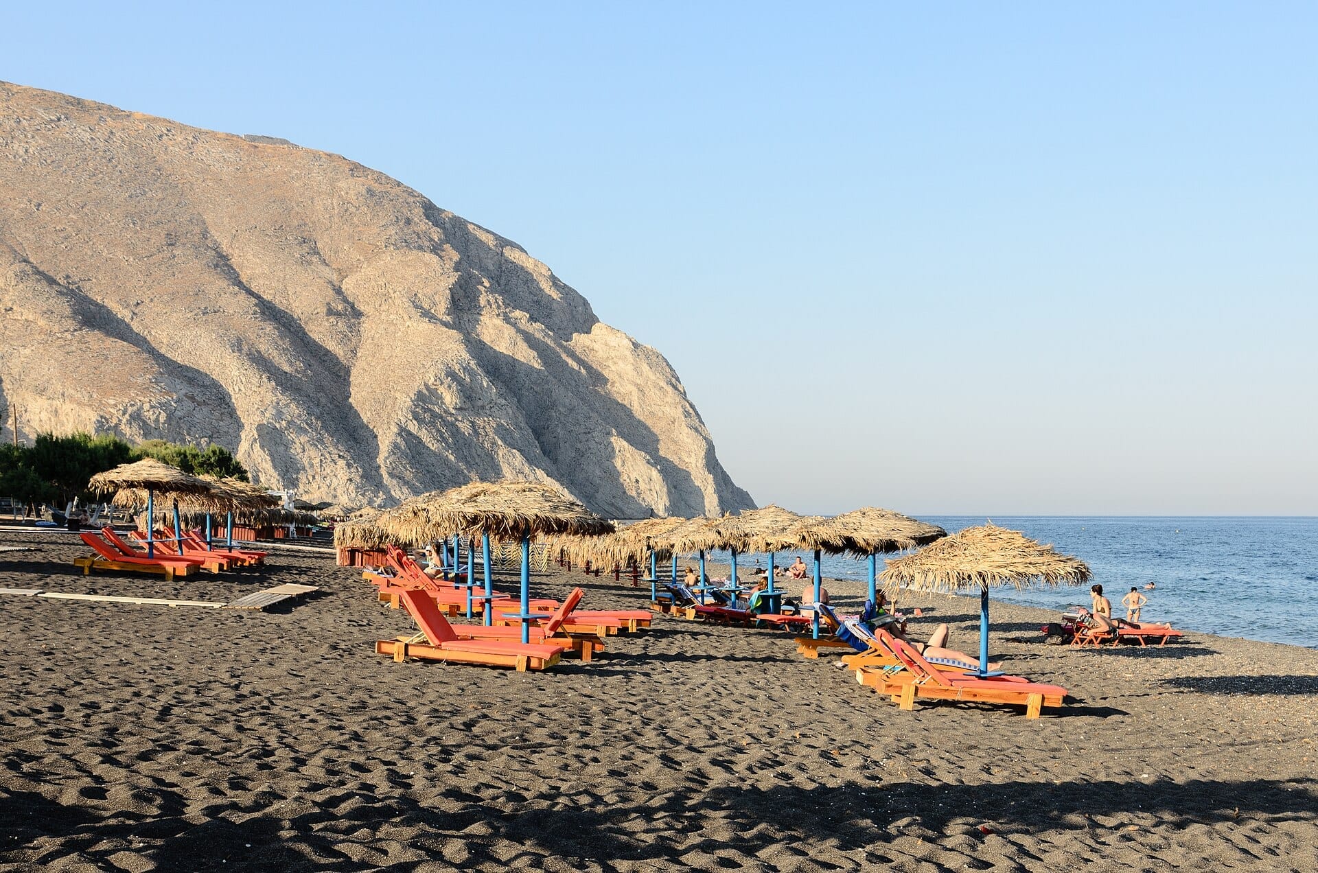 แถวเตียงอาบแดดและร่มฟางตั้งอยู่บนหาดทรายภูเขาไฟสีดำของ Perissa Beach โดยมี Mesa Vouno ตระหง่านอยู่เบื้องหลัง