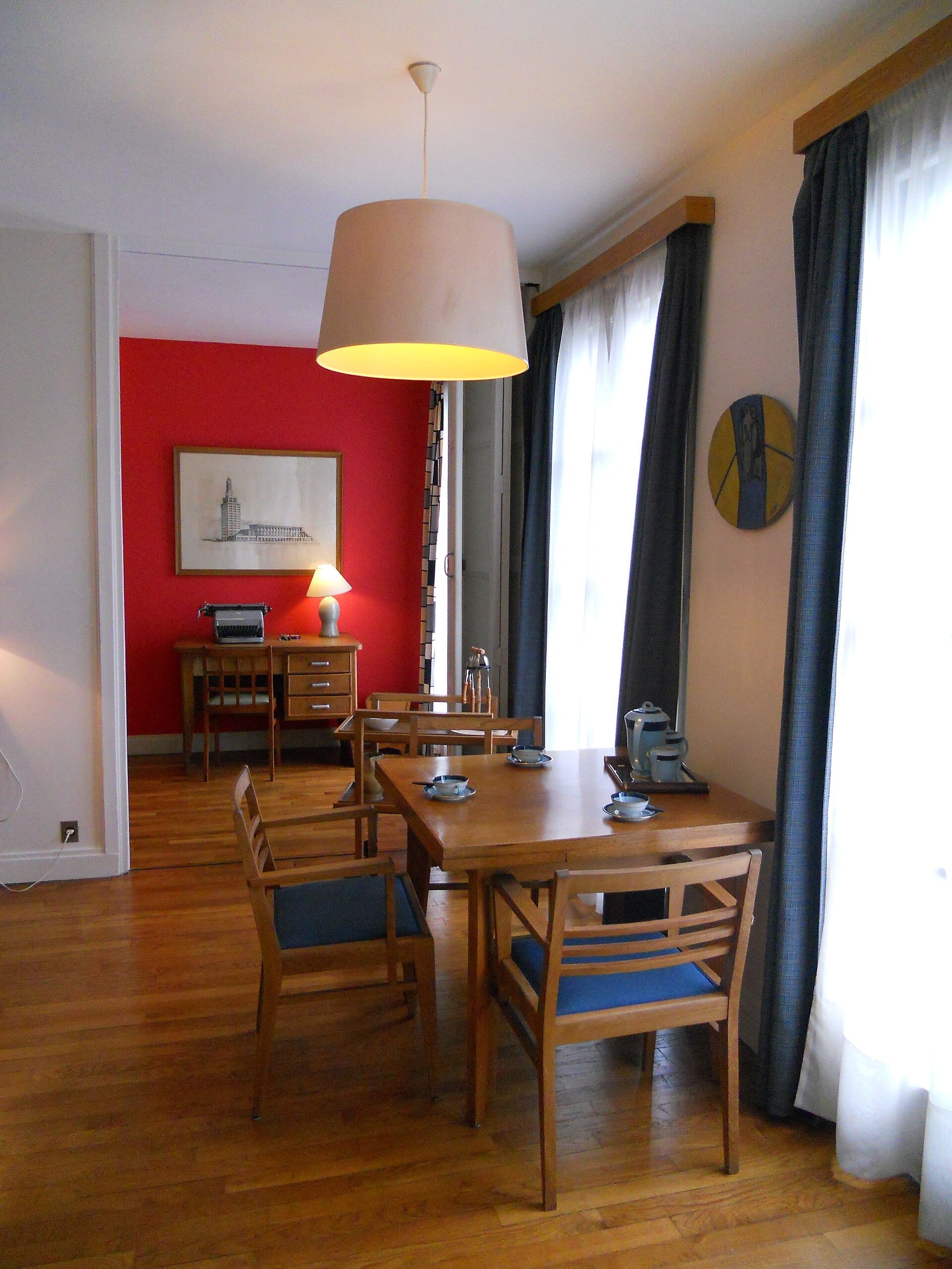 Sala da pranzo in stile moderno della metà del secolo nell'Appartement Témoin Perret, con mobili in legno, sedie blu, finestre luminose e una parete d'accento rossa.