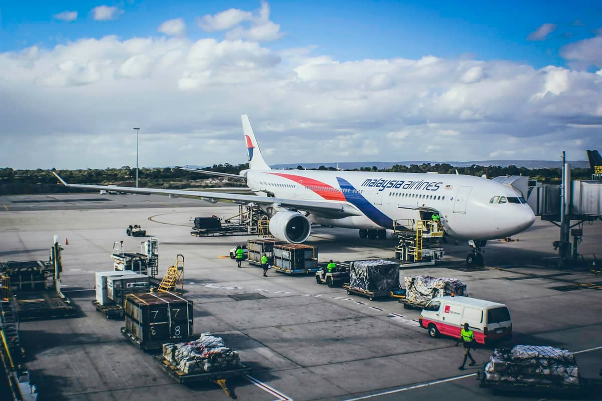 Malaysia Airlines aircraft parked at the gate at Perth Airport with baggage carts and ground crew visible under a partly cloudy sky.
