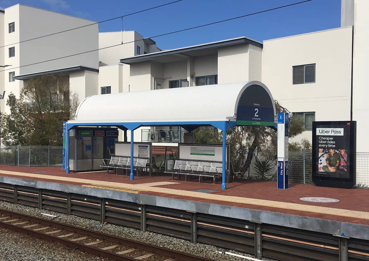 Typical Perth suburban train station platform with covered seating, ticket machine, and Uber Pass advertisement; apartment buildings visible in background.