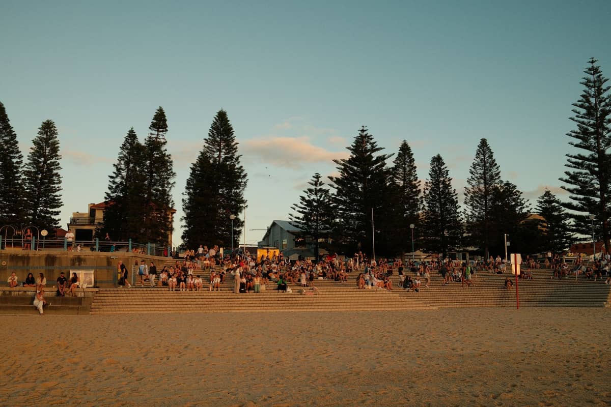 Menschenmassen versammeln sich auf breiten Sandstufen, gesäumt von hohen Norfolk-Kiefern, an einem klassischen Strand in Perth zur goldenen Stunde.