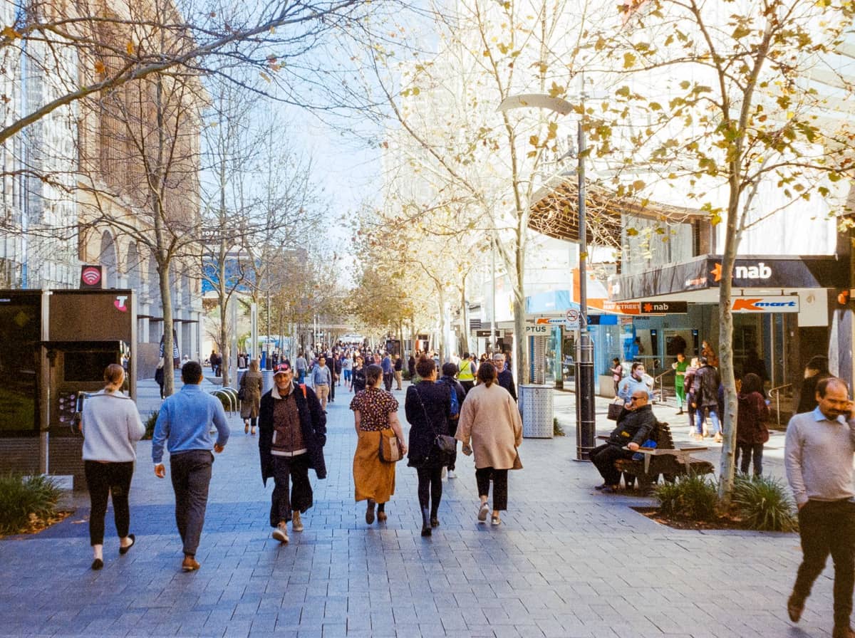 A busy pedestrian street in Perth with people walking past shops and cafes, trees lining the walkway, and a lively daytime atmosphere.