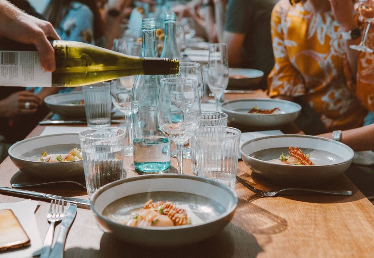 A long outdoor dining table set with plates of seafood, wine glasses, and people enjoying a meal together in natural daylight.