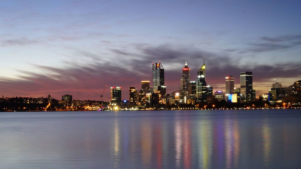 Perth city skyline at dusk with colorful lights reflecting on the calm water of the Swan River, under a dramatic evening sky.