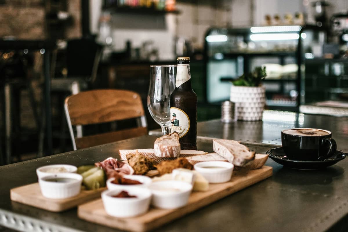 Charcuterie-Brett mit Brot, Käse, Aufschnitt und Beilagen auf einem Cafétisch mit einer Flasche Craft Beer und einer Kaffeetasse, gemütliches Innensetting.