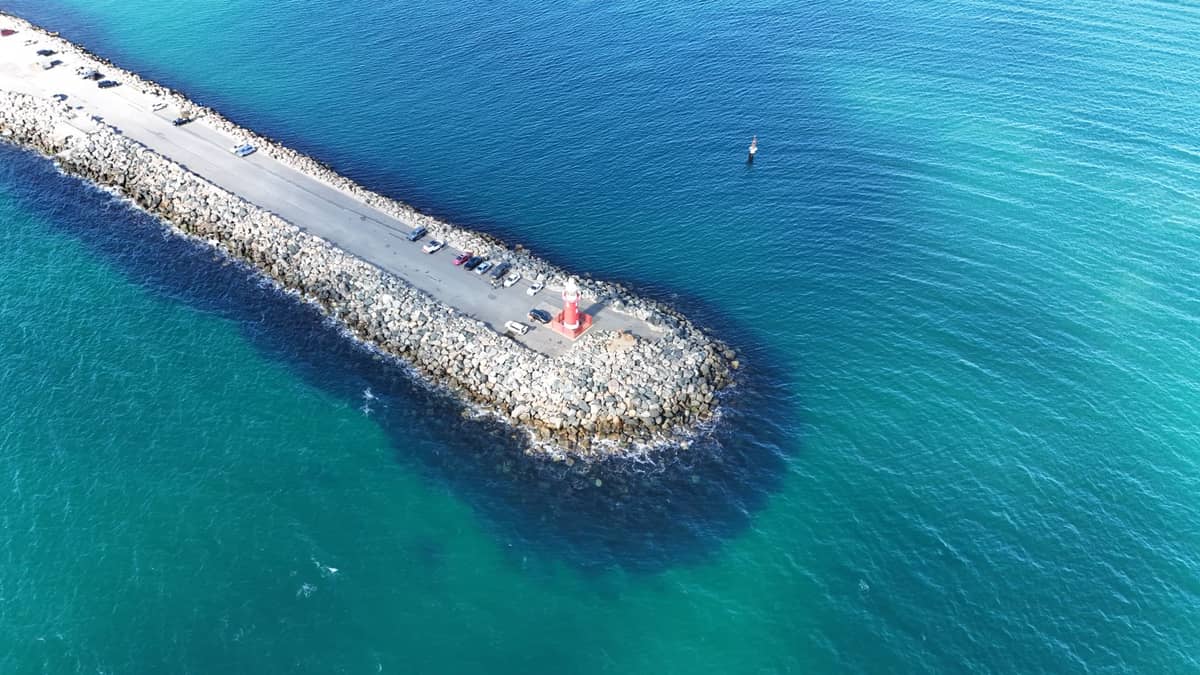 Widok z lotu ptaka na latarnię morską South Mole w Fremantle, stojącą na długim kamiennym molo wychodzącym w błękitne wody Oceanu Indyjskiego, z kilkoma osobami i samochodami w tle.