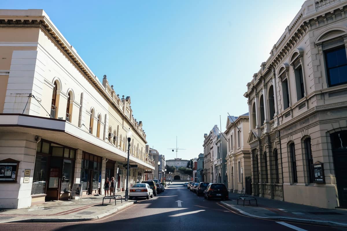Sonnige Straßenansicht in Fremantle mit historischen Gebäuden, breitem Gehsteig und klassischen Fassaden unter blauem Himmel, am Straßenrand geparkte Autos.
