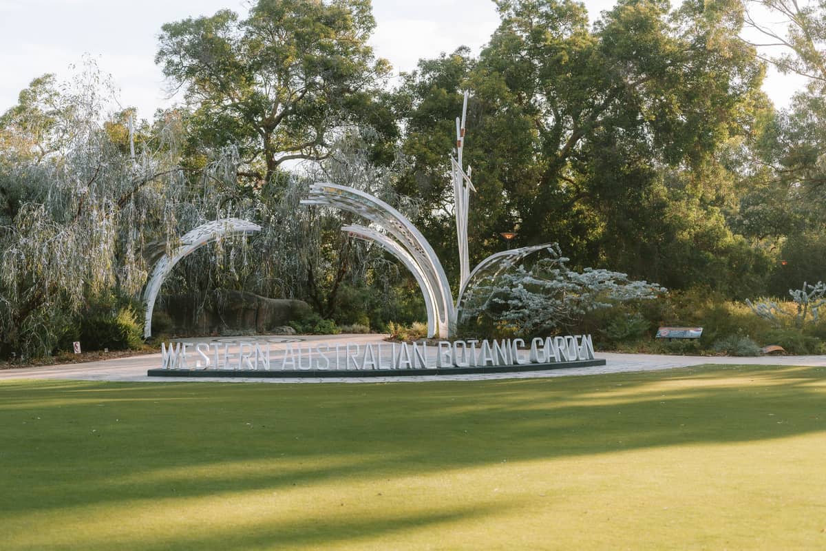 Eingangsschild des Western Australian Botanic Garden im Kings Park, umgeben von grünen Bäumen und Rasen unter klarem Himmel.