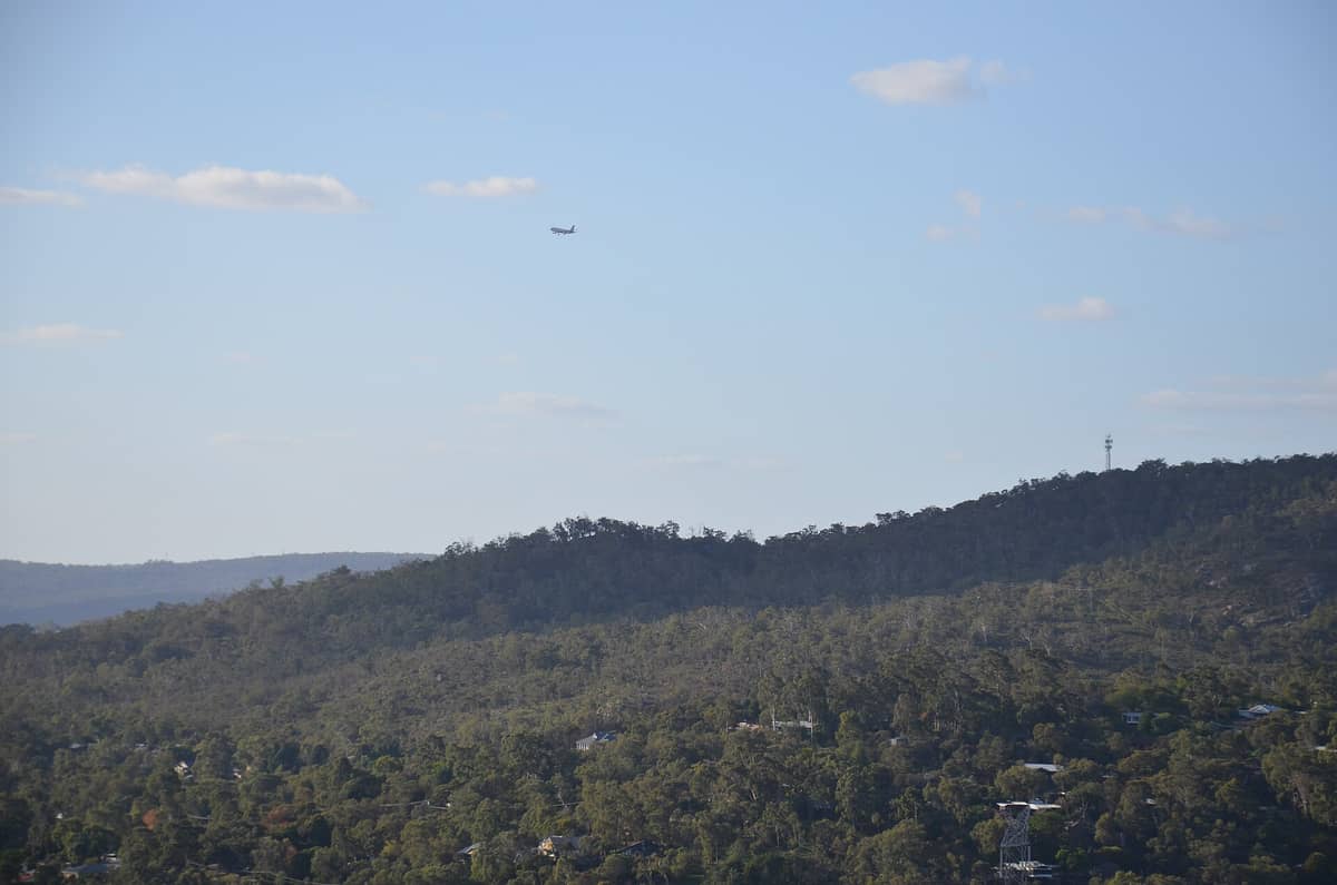 วิวมุมไกลของเนินภูเขาและต้นไม้นับไม่ถ้วนในภูมิภาค Perth Hills ภายใต้ท้องฟ้าสีฟ้าหม่น พร้อมเครื่องบินลำหนึ่งบินผ่านด้านบน