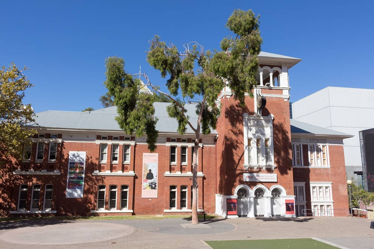 El exterior del Perth Institute of Contemporary Arts presenta un histórico edificio de ladrillo rojo con ventanas arqueadas, frondosos árboles y un cielo azul despejado.