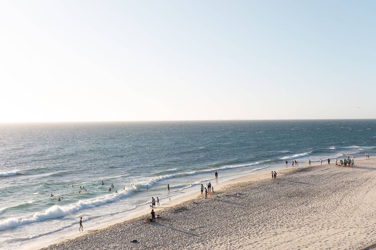 Weiter Sandstrand mit Menschen, die im sanften Wellengang spazieren und schwimmen, unter einem klaren Himmel, der die sommerliche Küstenatmosphäre von Perth heraufbeschwört.
