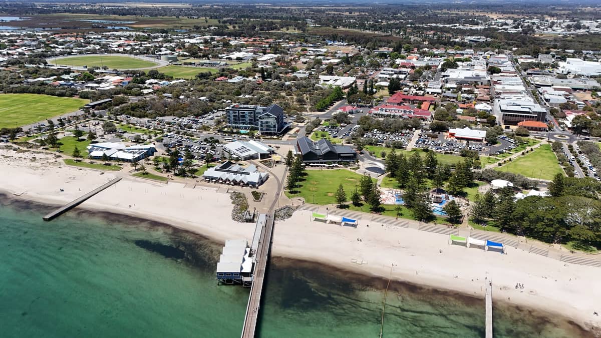Vista aérea de Bunbury, Australia Occidental, con el frente marítimo de la ciudad, espacios verdes y una playa de arena con un largo muelle que se extiende hacia aguas turquesas.