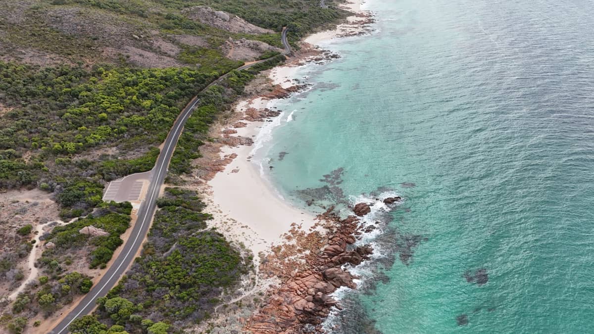 Una sinuosa carretera costera discurre junto a aguas turquesas y una orilla rocosa y arenosa bordeada de exuberante vegetación.