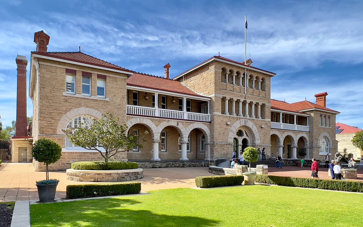 Weitwinkelansicht des historischen Sandsteingebäudes der Perth Mint mit markanten Bögen, rotem Dach, gepflegten Gärten und Besuchern am Haupteingang unter blauem Himmel.