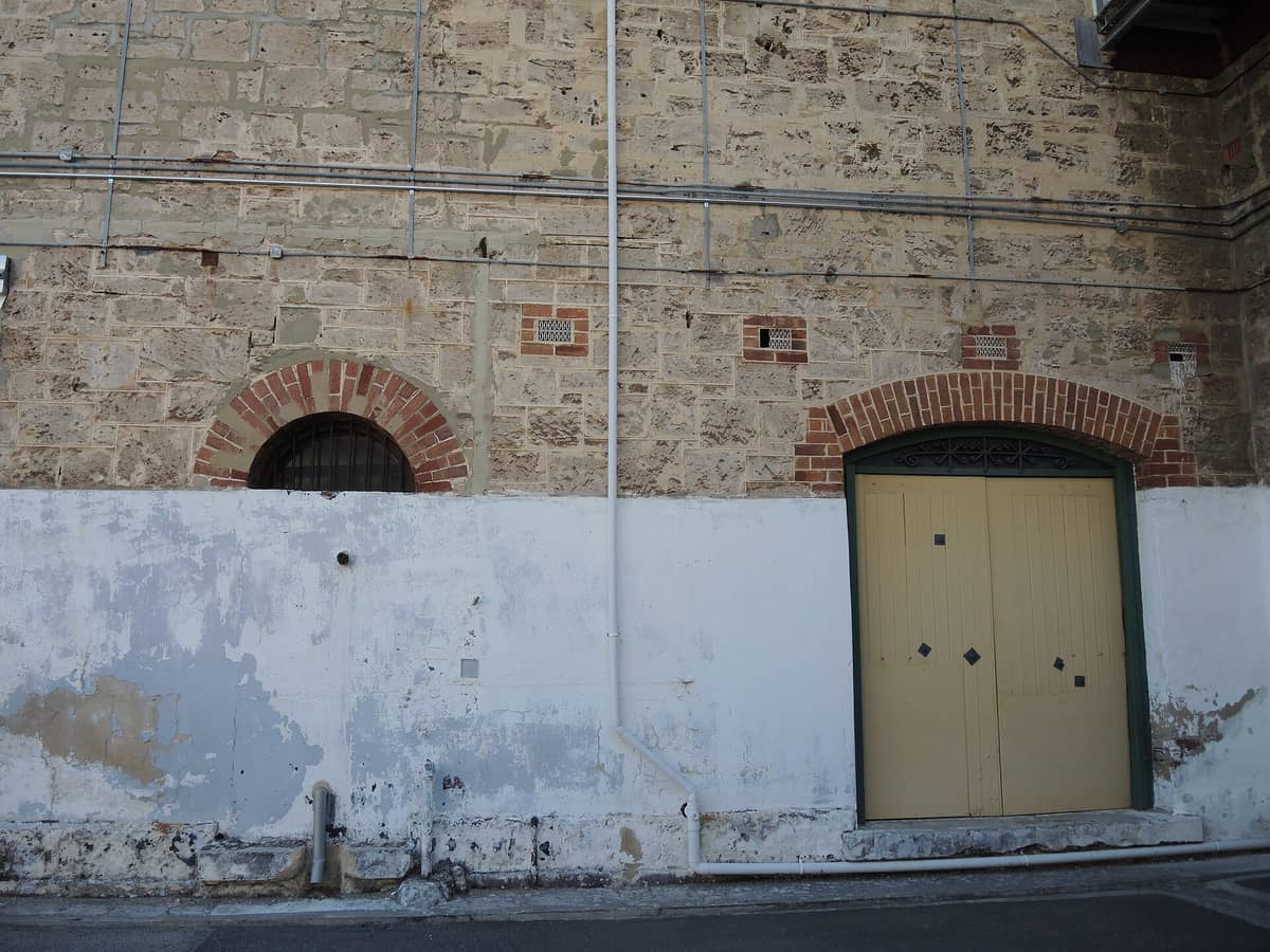 Außenwand der Fremantle Prison mit vergittertem Fenster und gewölbtem Eingang, mit verwittertem Stein und historischer Architektur