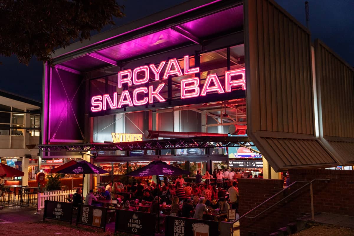 Belebte Nachtlebensszene vor der Royal Snack Bar mit Neonlichtern und Menschenmengen, die unter dem Abendhimmel an Tischen im Freien sitzen.